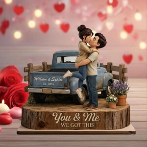 May include: A figurine of a couple kissing, standing in front of a blue pickup truck. The truck bed has the names "William & Sophia" and "Est. 2024". The base reads "You & Me WE GOT THIS". The figurine is on a wooden base.