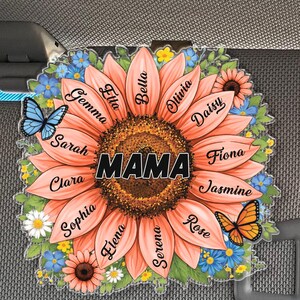 May include: A decorative sunflower-shaped ornament with the word "MAMA" in the center. The peach-colored petals are inscribed with names. Blue and yellow flowers and two butterflies surround the sunflower.