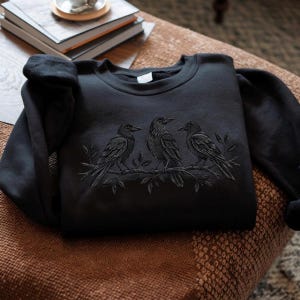 Puede incluir: Una sudadera negra de cuello redondo con un diseño bordado en tono sobre tono de tres cuervos posados en una rama con hojas. El bordado está realizado con un hilo oscuro que combina con la tela de la prenda, creando un efecto visual sutil y texturizado.