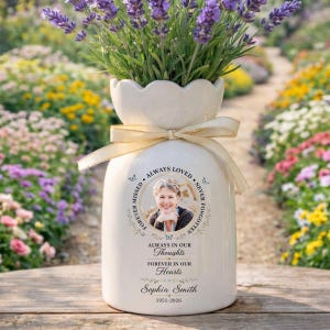 May include: A white ceramic vase with a cream-colored ribbon, holding lavender flowers. The vase features a photo and text that reads "Always Loved, Never Forgotten, Always in Our Thoughts, Forever in Our Hearts, Sophia Smith 1951-2026."