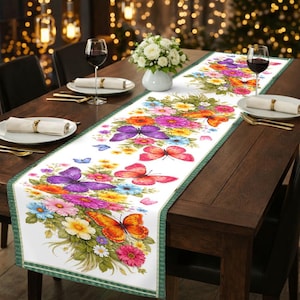 May include: A white table runner with a vibrant floral and butterfly design. The runner features colorful butterflies and flowers in shades of purple, orange, pink, and blue. It has a green border and is placed on a dark wooden table.