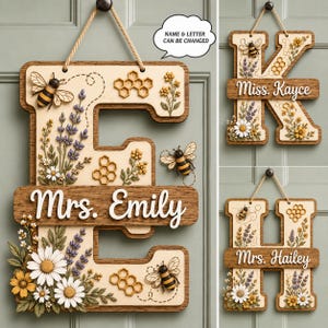 May include: A wooden wall hanging featuring a large letter E, K, or H with a natural wood border. The design includes raised 3D elements of bees, honeycombs, lavender, and daisies. A horizontal wooden plaque displays custom names like Mrs. Emily.