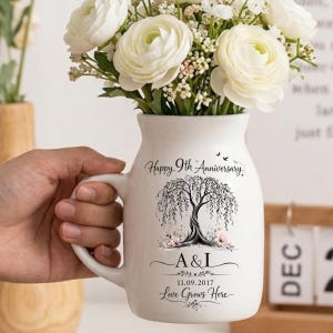 May include: White ceramic vase with a handle, filled with white flowers. The vase is decorated with a black and white weeping willow tree design and the text "Happy 9th Anniversary", "A & I", "11.09.2017", and "Love Grows Here".