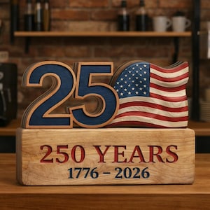 May include: Wooden commemorative display featuring the number "25" in blue, a waving American flag, and the text "250 YEARS 1776-2026" in red and blue. The display is made of wood and sits on a wooden surface.