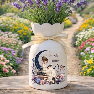 May include: White ceramic vase with a cream-colored bow, filled with lavender. The vase features a crescent moon design with a woman reading, the name "Sophia", and butterflies. The vase is decorated with flowers.