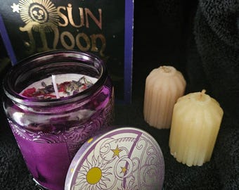 May include: A purple glass candle jar with a floral design and a lid featuring a sun and moon design with stars. The candle jar is sitting on a black surface next to two smaller white candles.