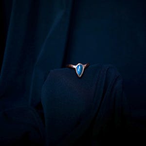 May include: A ring featuring a teardrop-shaped blue stone set in a metallic band. The stone displays a vibrant blue hue with a smooth, polished surface, while the band has a dark, textured finish that contrasts with the bright blue centre.