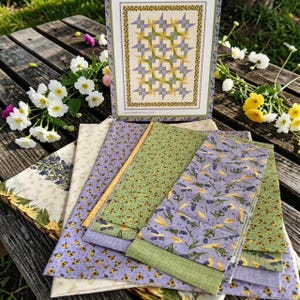 May include: A collection of floral fabric pieces in various patterns and colors, including lavender, green, and yellow. A framed quilt pattern with a star design is also present. The fabrics are arranged on a wooden surface.
