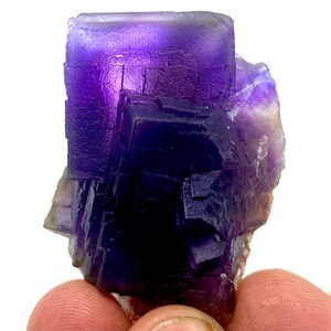May include: A vibrant purple fluorite crystal cluster with a cubic structure. The crystal displays a range of purple hues, from deep indigo to lighter shades, with a translucent quality. The edges of the crystal are well-defined, showcasing its natural formation.