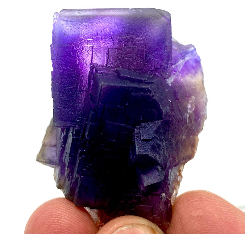 May include: A vibrant purple fluorite crystal cluster with a cubic structure. The crystal displays a range of purple hues, from deep indigo to lighter shades, with a translucent quality. The edges of the crystal are well-defined, showcasing its natural formation.