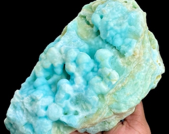 Op de afbeelding: Een groot, onregelmatig gevormd mineraal specimen met een opvallende, lichtblauwe kleur. Het oppervlak heeft een bubbelachtig, getextureerd uiterlijk, met gebieden in lichtere en donkerdere tinten blauw en vleugjes geelgroen. De achtergrond is zwart.