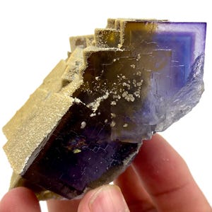 May include: A natural fluorite mineral specimen with a layered structure. The crystal features a gradient of colors, including deep purple, yellow, and tan. The surface has a textured appearance with small, white crystalline deposits.