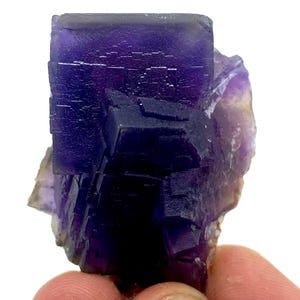 May include: A close-up of a purple fluorite crystal cluster. The crystal has a cubic structure with a deep purple color, transitioning to lighter shades. The edges of the crystal are well-defined, and the surface has a textured appearance.
