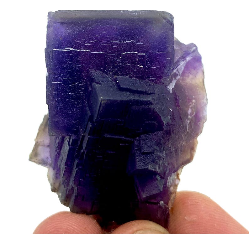 May include: A close-up of a purple fluorite crystal cluster. The crystal has a cubic structure with a deep purple color, transitioning to lighter shades. The edges of the crystal are well-defined, and the surface has a textured appearance.
