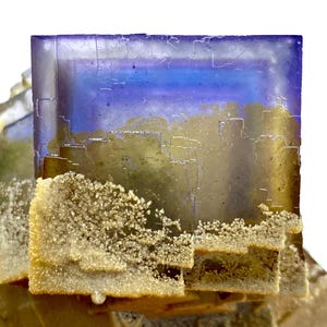 May include: A close-up of a fluorite crystal with a layered, geometric structure. The crystal features a gradient of colors, including blue, purple, and gold. The lower portion has a textured, crystalline coating, adding contrast to the smooth surfaces.