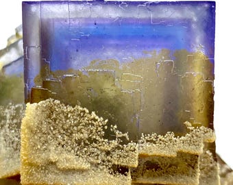 May include: A close-up of a fluorite crystal with a layered, geometric structure. The crystal features a gradient of colors, including blue, purple, and gold. The lower portion has a textured, crystalline coating, adding contrast to the smooth surfaces.