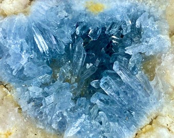 May include: A cluster of translucent, light blue celestite crystals forming a jagged, geometric formation. The central blue crystals are surrounded by a rough, textured border of pale, off-white mineral deposits with a small yellow spot.