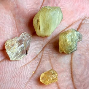 以下が含まれることがあります： 黄色と透明の4つの原石。石は不規則な形で、インクルージョンとテクスチャが見えます。宝石は、おそらく手であるピンク色の背景に表示されています。