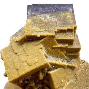 May include: Close-up of a mineral specimen featuring a cluster of cubic and layered formations. The mineral has a light tan color with a crystalline texture, and a dark purple, almost black, section at the top. The background is white.