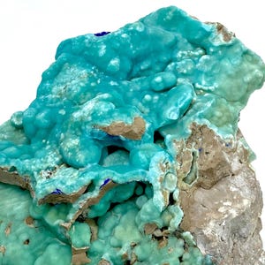 May include: A close-up of a vibrant turquoise mineral specimen. The mineral has a bubbly, textured surface with areas of light brown and gray rock. Small patches of deep blue are visible throughout the specimen.
