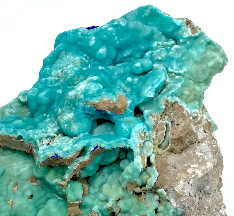 May include: A close-up of a vibrant turquoise mineral specimen. The mineral has a bubbly, textured surface with areas of light brown and gray rock. Small patches of deep blue are visible throughout the specimen.