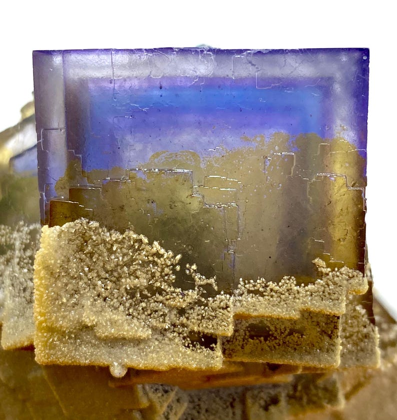 May include: Close-up of a fluorite crystal formation. The crystal features a gradient of colors, including purple, blue, and gold. The lower portion of the crystal has a textured, crystalline appearance. The background is white.