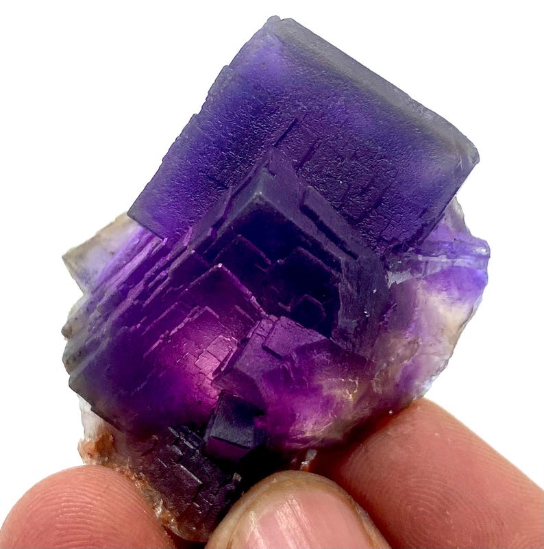 May include: A close-up of a vibrant purple fluorite crystal cluster. The crystal exhibits a cubic structure with sharp edges and a deep, saturated color gradient. The base of the crystal is a light brown color.