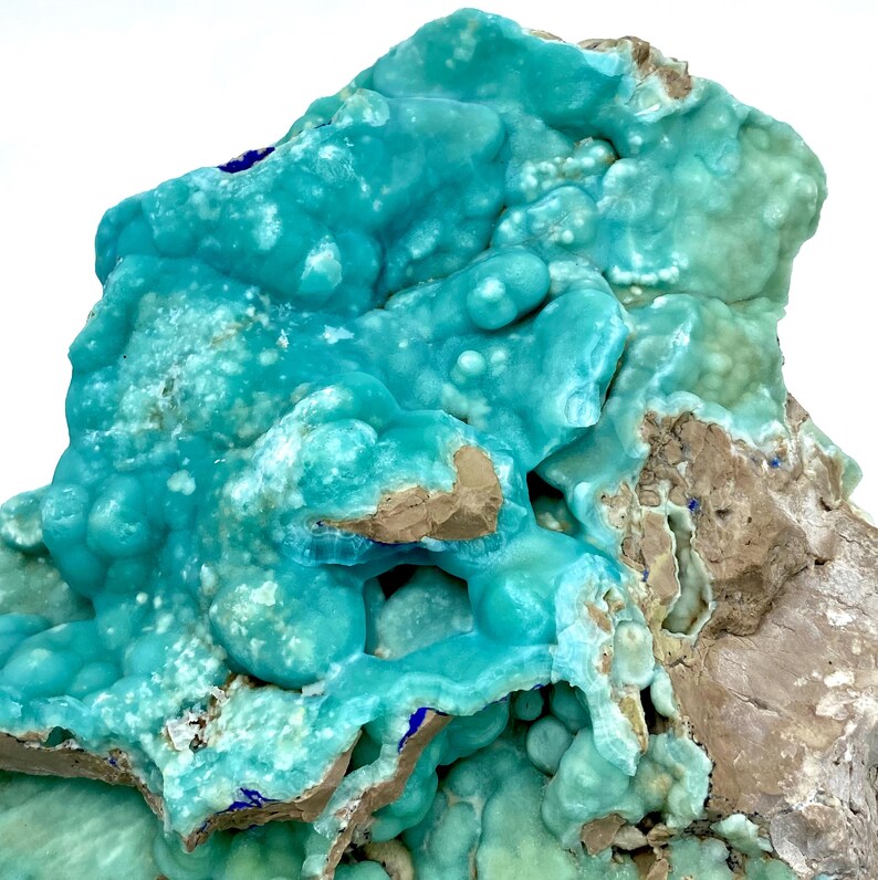 May include: A close-up of a mineral specimen featuring a vibrant turquoise hue with a bubbly, textured surface. The mineral has areas of light brown and gray rock, with small patches of dark blue.