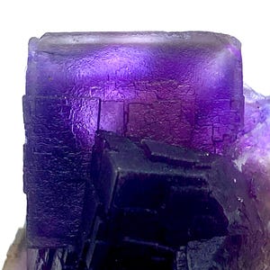 May include: Close-up of a purple fluorite crystal formation. The crystal has a cube-like structure with a textured surface and varying shades of purple. The crystal is set against a white background.