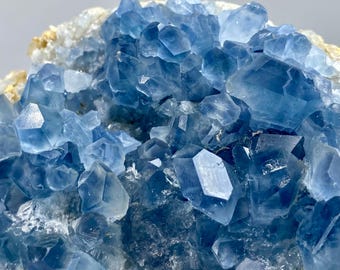 May include: Close-up of a cluster of blue celestite crystals. The crystals are a light, translucent blue with sharp, geometric shapes. The base of the cluster is a mix of white and tan. The overall effect is a display of natural mineral beauty.