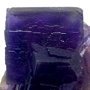 May include: Close-up of a vibrant purple fluorite crystal formation. The crystal exhibits a cubic structure with a deep, saturated color. The surface has a textured appearance, with visible crystalline details. The overall presentation is a natural, geological specimen.