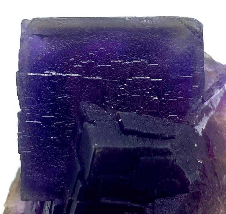 May include: Close-up of a vibrant purple fluorite crystal formation. The crystal exhibits a cubic structure with a deep, saturated color. The surface has a textured appearance, with visible crystalline details. The overall presentation is a natural, geological specimen.
