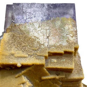 May include: Close-up of a natural fluorite mineral specimen. The top portion is a deep purple color, transitioning to a golden-yellow hue with a crystalline texture. The specimen has a layered, geometric structure.