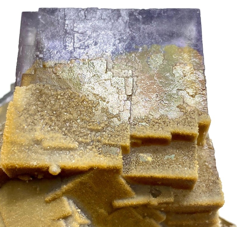 May include: Close-up of a natural fluorite mineral specimen. The top portion is a deep purple color, transitioning to a golden-yellow hue with a crystalline texture. The specimen has a layered, geometric structure.