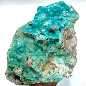 May include: A vibrant, natural mineral specimen featuring a striking combination of colors. The piece showcases a textured, turquoise-blue surface with areas of lighter green and tan hues. The overall form is irregular and organic, highlighting its raw, unrefined beauty.