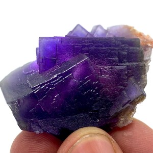 May include: A close-up of a fluorite mineral specimen. The fluorite features deep purple and violet cubic crystals. The specimen has a rough, textured appearance with a small amount of tan-colored matrix.