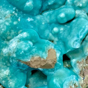 May include: Close-up of a mineral specimen with a striking turquoise hue. The surface has a bubbly, textured appearance with areas of lighter blue and patches of tan-colored rock. The overall effect is reminiscent of a frozen wave.