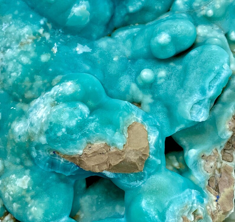 May include: Close-up of a mineral specimen with a striking turquoise hue. The surface has a bubbly, textured appearance with areas of lighter blue and patches of tan-colored rock. The overall effect is reminiscent of a frozen wave.