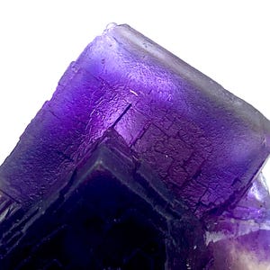 May include: Close-up of a vibrant purple fluorite crystal formation. The crystal exhibits a layered, geometric structure with varying shades of purple, from deep violet to lighter hues. The surface has a textured appearance, with visible crystalline patterns.