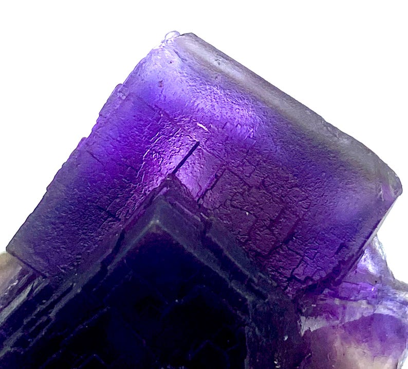 May include: Close-up of a vibrant purple fluorite crystal formation. The crystal exhibits a layered, geometric structure with varying shades of purple, from deep violet to lighter hues. The surface has a textured appearance, with visible crystalline patterns.