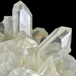 May include: A cluster of clear, translucent quartz crystals featuring sharp, pointed terminations and multifaceted surfaces. The mineral formation displays varying degrees of transparency and internal inclusions, set against a solid black background.