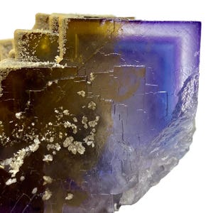 May include: Close-up of a fluorite crystal with a layered, geometric structure. The crystal displays a gradient of colors, including deep purple, blue, and golden yellow. Small, white crystalline formations are scattered across the surface, adding texture.