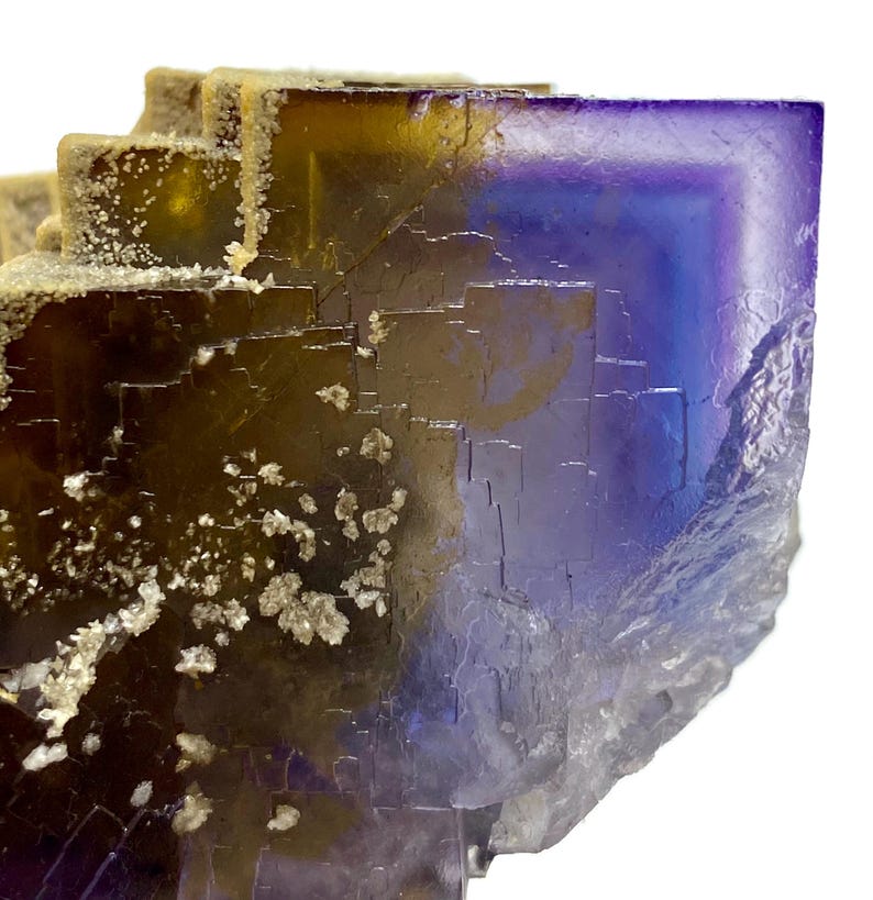 May include: Close-up of a fluorite crystal with a layered, geometric structure. The crystal displays a gradient of colors, including deep purple, blue, and golden yellow. Small, white crystalline formations are scattered across the surface, adding texture.