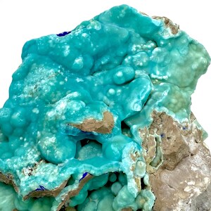 May include: A close-up of a mineral specimen featuring a vibrant turquoise hue. The surface is textured with rounded formations and patches of brown and gray rock. Small blue accents are visible, creating a striking contrast. The overall appearance suggests a natural, geological formation.
