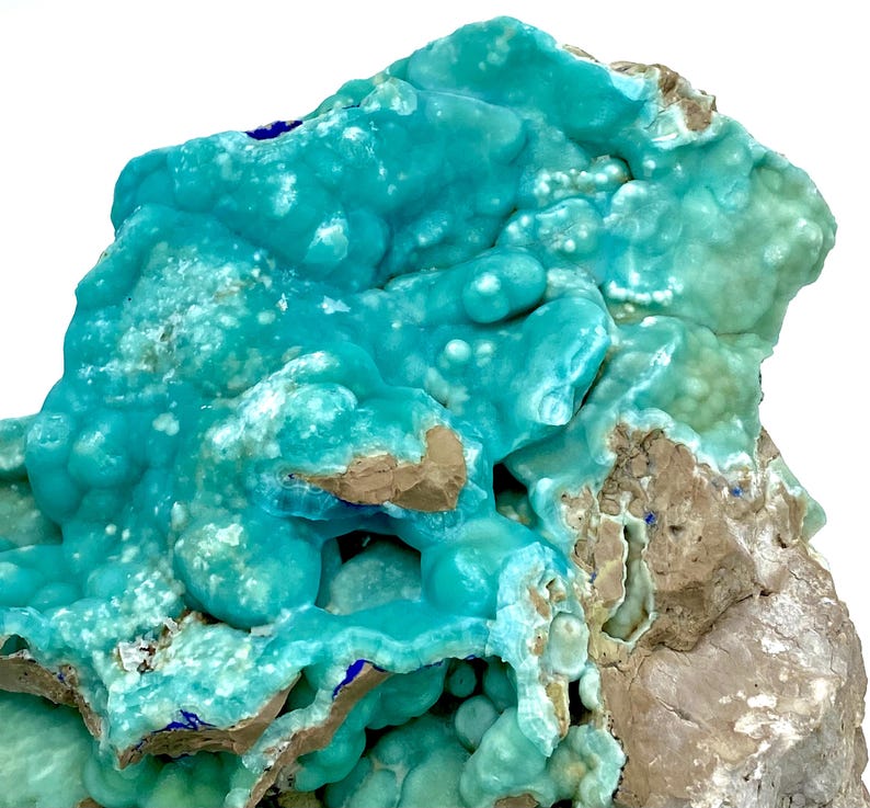 May include: A close-up of a mineral specimen featuring a vibrant turquoise hue. The surface is textured with rounded formations and patches of brown and gray rock. Small blue accents are visible, creating a striking contrast. The overall appearance suggests a natural, geological formation.