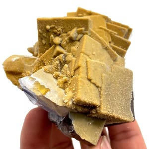 May include: A close-up of a mineral specimen featuring a cluster of golden-brown, cubic crystals. The crystals have a rough, textured surface and are arranged in a layered formation. The base of the specimen shows hints of purple and white.