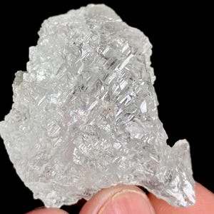 Puede incluir: Un espécimen mineral claro y cristalino con una superficie rugosa y texturizada. La piedra translúcida tiene un patrón geométrico y una ligera iridiscencia arcoíris. El fondo es negro, resaltando la claridad y la forma del mineral.
