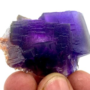 May include: A close-up of a vibrant purple fluorite crystal cluster. The crystal exhibits a deep, rich color with a complex, geometric structure. The edges of the crystal are sharp and well-defined, showcasing its natural formation. The crystal is held by a person's fingers.