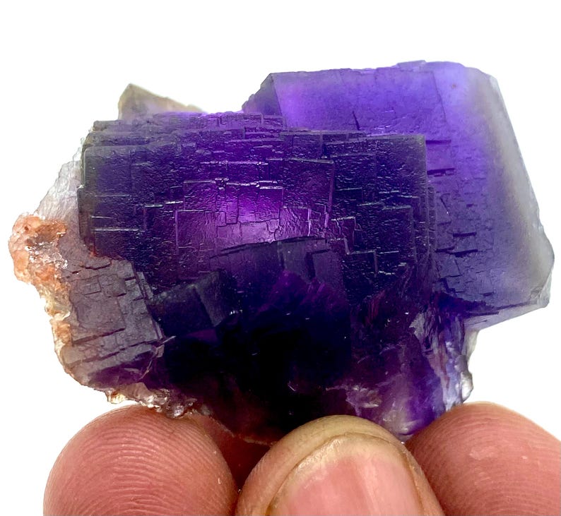 May include: A close-up of a vibrant purple fluorite crystal cluster. The crystal exhibits a deep, rich color with a complex, geometric structure. The edges of the crystal are sharp and well-defined, showcasing its natural formation. The crystal is held by a person's fingers.