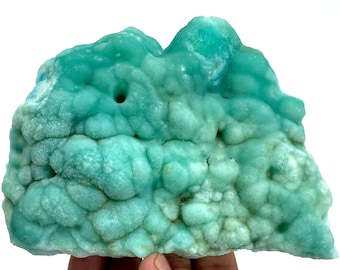 May include: A vibrant, turquoise-colored mineral specimen with a bubbly, textured surface. The stone has a unique, organic shape and a few small holes. The mineral is held by a hand, showcasing its size and color against a white background.